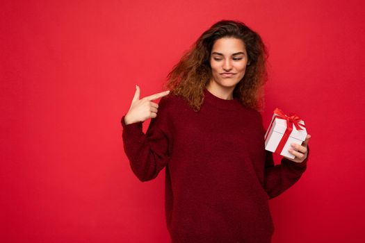 Attractive positive self-confident emotional young brunette curly woman isolated over red background wall wearing red sweater holding gift box looking to the side.