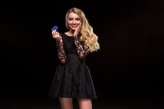 Beautiful blonde in a black dress with casino chips in hands isolated on a black background. Poker. Casino. Roulette Blackjack Spin. Caucasian young woman looking at the camera