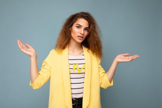 Photo shot of young asking thinking nice cute brunette curly woman with sincere emotions wearing trendy yellow jacket isolated on blue background with copy space and having doubts.