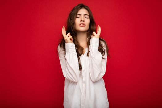 Shot of attractive young curly brunette woman wearing white shirt isolated on red background with copy space, praying and dreaming.