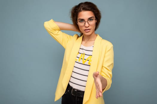 Photo of young self-confident attractive brunette curly lady with sincere emotions wearing stylish yellow jacket and optical glasses isolated on blue background with copy space.