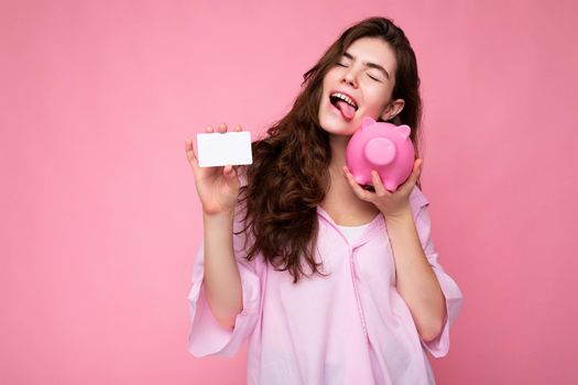 Attractive happy young brunette woman wearing shirt isolated on pink background with empty space and holding pink pig money box and credit card for mockup.