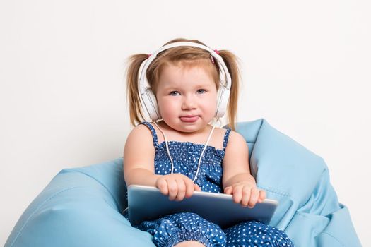 Cute little girl in headphones listening to music using a tablet and smiling while sitting on blue big bag. On white background. A child looks at the camera