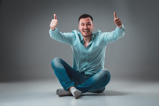 Handsome young man sitting on a floor with raised hands, isolated on gray background. Emotion concept