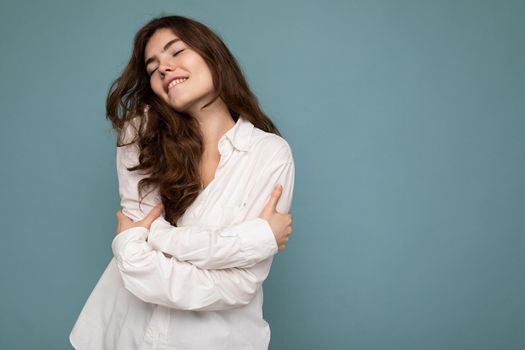 Portrait of young positive happy satisfied attractive curly brunette woman with sincere emotions wearing casual white shirt isolated on blue background with empty space and hugging herself.