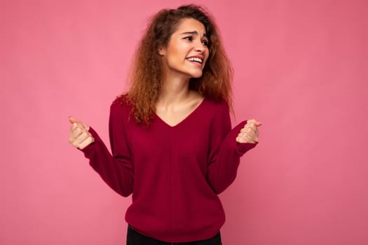Photo of young emotional positive happy pretty brunette curly woman with sincere emotions wearing casual pink sweater isolated over pink background with copy space.