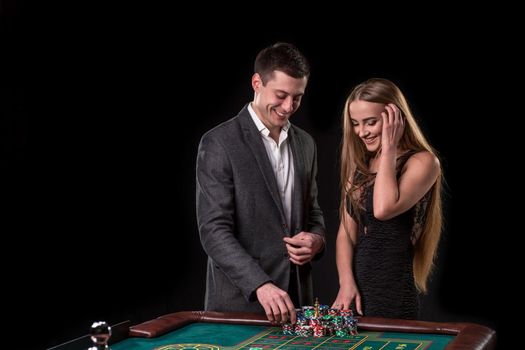 Elegant couple at the casino betting on the roulette, on a black background. A man in a suit with a beautiful young woman in a black dress