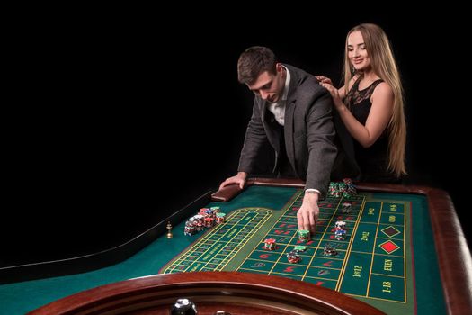 Elegant couple at the casino betting on the roulette, on a black background. A man in a suit with a beautiful young woman in a black dress