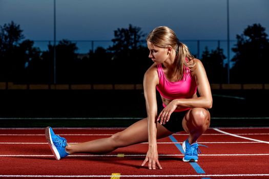 Attractive young woman athlete stretching legs on stadium. The end of training at sunset.