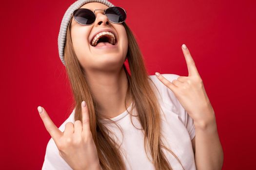 Closeup of young emotional positive happy attractive dark blonde woman with sincere emotions wearing casual white t-shirt, gray hat and sunglasses isolated on red background with copy space and showing rock and roll gesture.