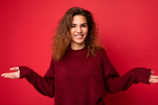 Young charming curly brunette female person with sincere emotions poising isolated on background wall with copy space wearing casual dark red sweater. Happy concept.