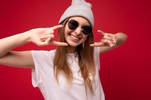 Closeup of young emotional positive happy smiling attractive dark blonde woman with sincere emotions wearing casual white t-shirt with empty space for mockup gray hat and sunglasses isolated on red background with copy space and showing rock and roll gesture.