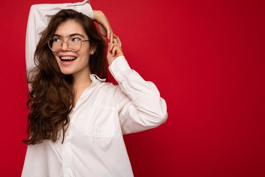 Photo of young beautiful happy smiling brunette woman wearing white shirt and optical glasses. Sexy carefree female person posing isolated near red wall in studio with free space. Positive model with natural makeup.