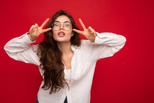 Shot of attractive positive happy joyful funny young curly brunette woman wearing white shirt and optical glasses isolated on red background with free space and showing peace gesture. Emotional concept.