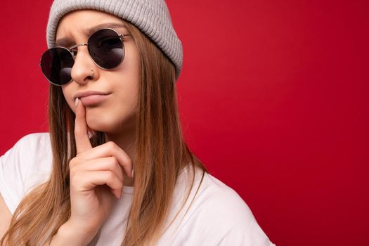 Closeup of adult emotional thinking dreaming beautiful dark blond woman with sincere emotions wearing casual white t-shirt, gray hat and sunglasses isolated on red background with copy space.
