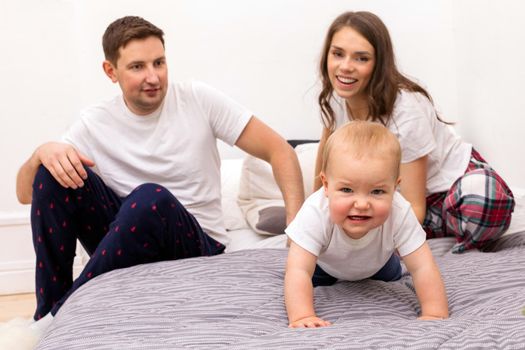 Little toddler standing on all fours on bed and looking at camera with satisfied young father and mother on background