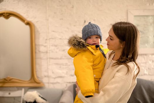 Little kid in yellow warm jacket and hat looking at camera in arms of young mother at home