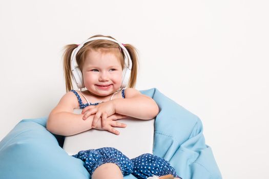 Cute little girl in headphones listening to music using a tablet and smiling while sitting on blue big bag. On white background. A child looks at the camera