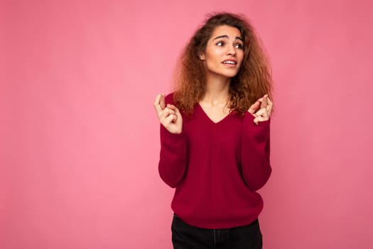 Photo of young emotional attractive brunette curly woman with sincere emotions wearing trendy pink sweater isolated over pink background with empty space and holding fingers crossed for good luck. Gesture concept.