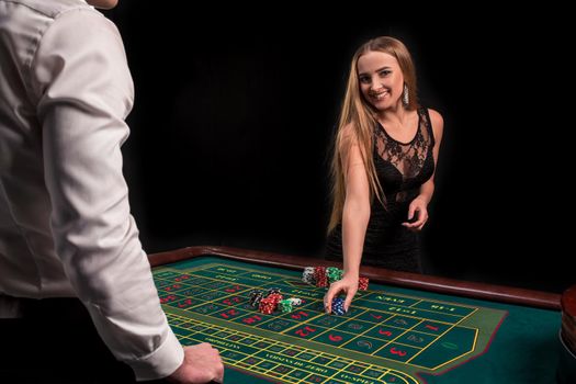 A close-up on the back of the croupier in a white shirt, image of green casino table with roulette and chips, a rich woman betting of gambling in the background. Casino. Gambling. Roulette. Betting