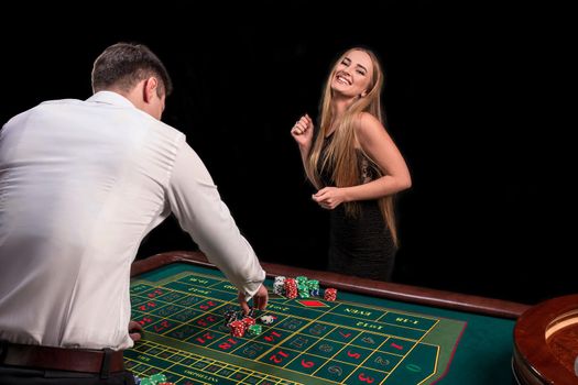 A close-up on the back of the croupier in a white shirt, image of green casino table with roulette and chips, a rich woman betting of gambling in the background. Casino. Gambling. Roulette. Betting