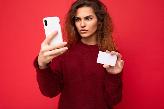 Young dissatisfied beautiful curly brunette woman with sincere emotions poising isolated over background wall with empty space wearing casual dark red sweater holding credit card and using mobile phone . Problems with Internet and payments.
