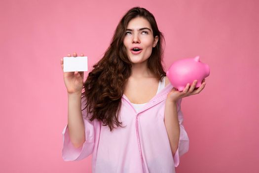 Attractive happy surprised young brunette woman wearing shirt isolated on pink background with empty space and holding pink pig money box and credit card for mockup.
