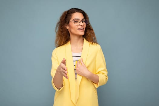 Photo of young positive happy attractive brunette curly woman with sincere emotions wearing stylish yellow jacket and optical glasses isolated on blue background with copy space.