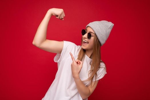 Portrait of young emotional positive happy beautiful dark blonde woman with sincere emotions wearing casual white t-shirt with empty space for mockup gray hat and sunglasses isolated over red background with free space. Female model showing powerful muscles and biceps. Fitness and workout concept.