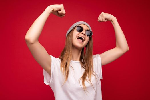 Portrait of young emotional positive happy attractive dark blonde woman with sincere emotions wearing casual white t-shirt with empty space for mockup grey hat and sunglasses isolated over red background with copy space and showing muscles. Workout sport motivation lifestyle concept.