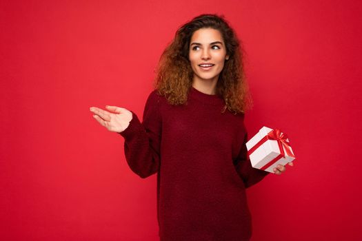 Beautiful happy young brunette woman isolated over colourful background wall wearing stylish casual clothes holding gift box and looking to the side.