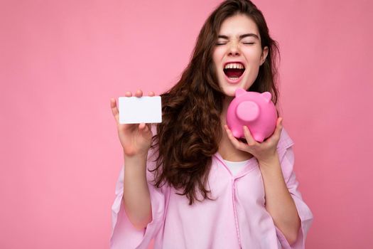 Attractive happy emotional young brunette woman wearing shirt isolated on pink background with empty space and holding pink pig money box and credit card for mockup.