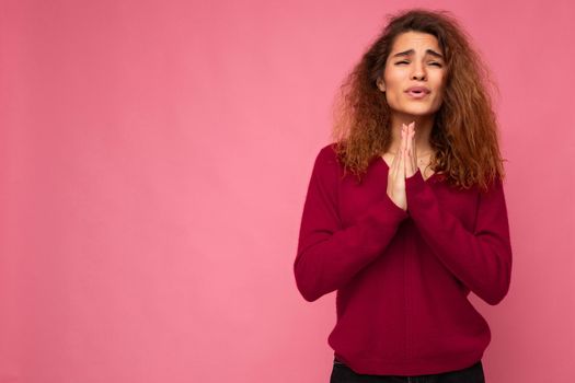 Photo of young emotional beautiful brunette curly woman with sincere emotions wearing trendy pink pullover isolated over pink background with free space and keeping hands in praying gesture and believing in good luck.