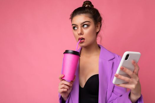 Shot of thoughtful caucasian brunette woman holds take away coffee dressed in casual clothes isolated over pink wall uses smartphone and drinking. Empty space