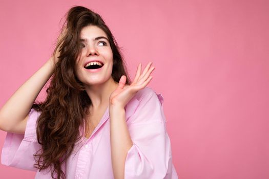 Photo of young positive happy adorable beautiful wavy-haired brunette woman with sincere emotions wearing trendy pink shirt isolated over pink background with free space.