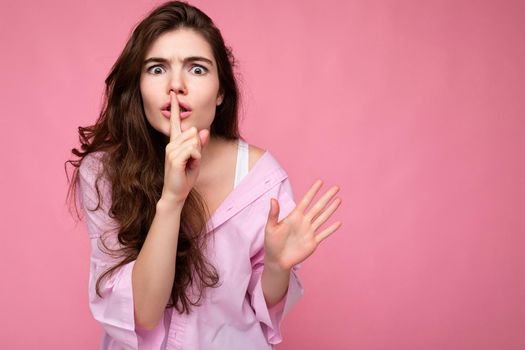 Photo of young emotional nervous beautiful wavy-haired brunette woman with sincere emotions wearing casual pink shirt isolated over pink background with copy space and showing silence gesture with finger near lips. Shhh sign.