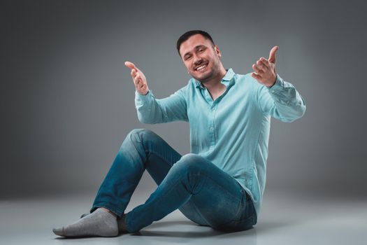 Handsome young man sitting on a floor with raised hands gesturing happiness on gray background. A man in jeans and a blue shirt