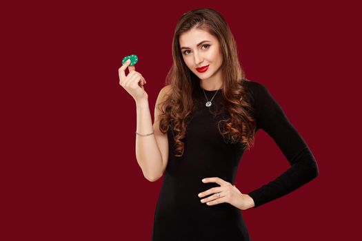 Pretty long hair woman in black dress holding chips for gambling in casino. Studio shot on red background. Poker. Two green Chips in Hand