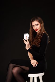 Sexy curly hair brunette in black dress posing with two aces cards in her hands, poker concept on black background. winning combination. sits on a chair