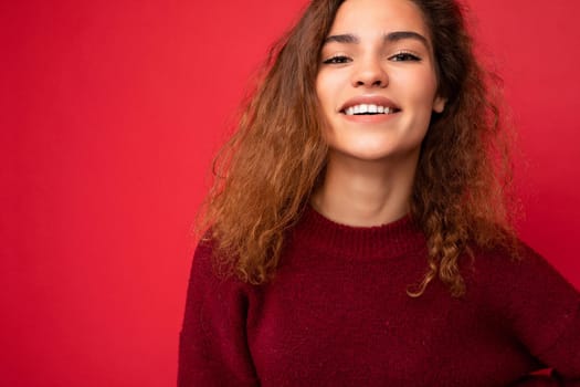 Young beautiful curly brunette woman with sincere emotions poising isolated on background wall with copy space wearing casual dark red sweater. Joy concept.