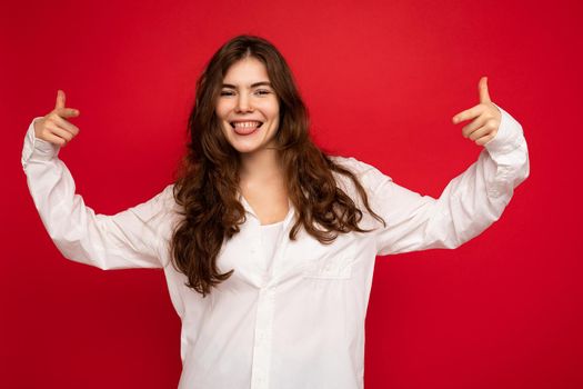 Photo of young beautiful happy smiling brunette woman wearing white shirt. Sexy carefree female person posing isolated near red wall in studio with free space. Positive model with natural makeup.