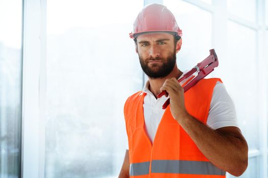 Young male plumber in workwear holding pipe wrench on a construction site indoors