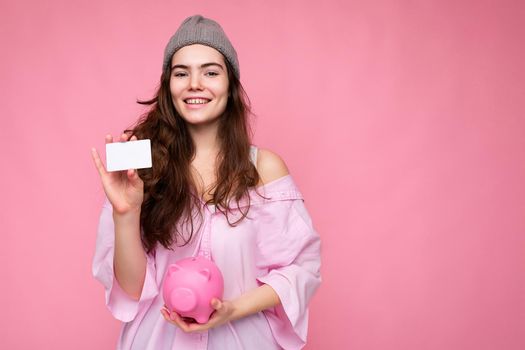 Portrait of beautiful positive cheerful cute smiling young brunette woman in stylish shirt isolated on pink background with copy space and holding pink pig moneybox and credit card for mockup.