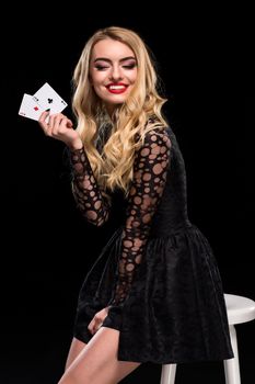 Beautiful confident woman in black dress sitting on chair showing cards looking at camera, isolated on the black background. With copy place Poker. Casino. Roulette Blackjack Spin.