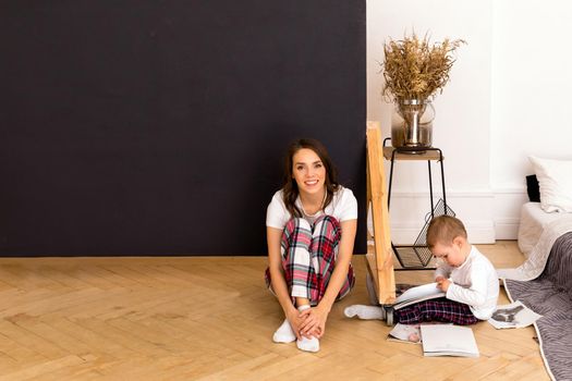Calm child drawing on paper sitting on floor with bright charming mom with crossed legs at home looking at camera