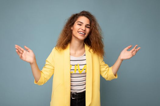Photo shot of young smiling positive happy attractive brunette curly woman with sincere emotions wearing trendy yellow jacket isolated on blue background with empty space and having doubts.