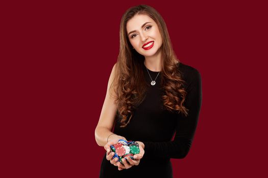 Casino, gambling, poker, people and entertainment concept - woman poker player in black dress with chips in hands on red background. Studio shot