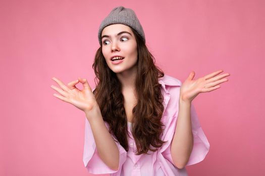 Beautiful strange funny young curly brunette woman wearing pink shirt and grey hat isolated on pink background with copy space and don't understanding.
