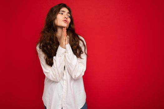 Portrait of beautiful young curly brunette woman wearing white shirt isolated on red background with copy space, praying and dreaming with sincere emotions.