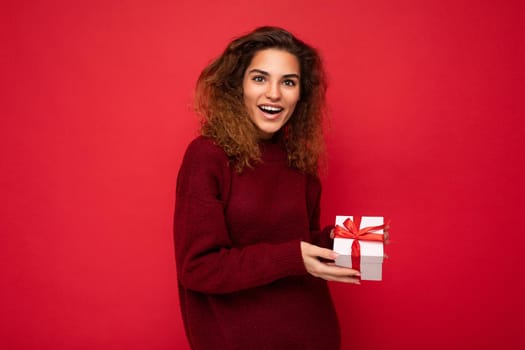 Attractive positive surprised young brunette curly woman isolated over red background wall wearing red sweater holding gift box looking at camera.